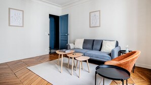 A modern living room with a grey sofa, two side tables, a black chair, and a woven ceiling light. - Blueground | Ternes, nr restaurants & attractions (Paris)