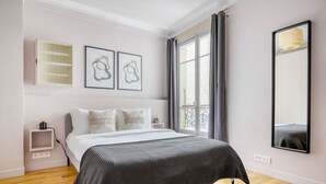A bedroom with a bed, a bedside table, a nightstand, a window with curtains, and a wall-mounted mirror. - Blueground | Ternes, nr restaurants & attractions (Paris)