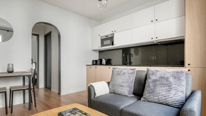 A modern living room with a grey sofa, a coffee table with a book, a kitchen area with white cabinets and a microwave, and a wooden floor.