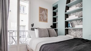 A bedroom with a bed, a balcony, a wall-mounted shelf with towels, and a framed picture.