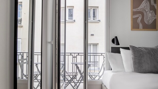 A hotel room with a bed, a bedside table, a lamp, and a view of the balcony with outdoor furniture.