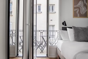 A hotel room with a bed, a bedside table, a lamp, and a view of the balcony with outdoor furniture. - Blueground | Champ de Mars, furnished, balcony (Paris)