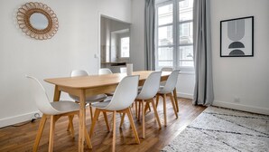 Une salle à manger moderne avec une table en bois et des chaises blanches, une grande fenêtre avec des rideaux et une œuvre d’art encadrée au mur.