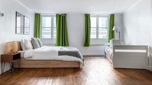 Une chambre avec un plancher en bois, un lit avec une literie blanche, deux fenêtres avec des rideaux verts, une table de chevet avec une lampe et une commode blanche.