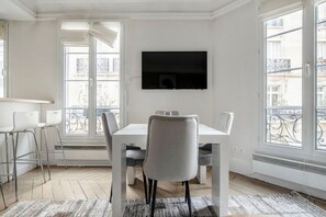 A modern dining area with a white table and chairs, a flat-screen TV mounted on the wall, and large windows with a view of the city. - Blueground | Trocadero, nr metro & parks (Paris)