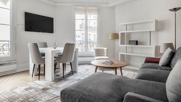 A modern living room with a dining area, a grey sofa, a coffee table, and a TV mounted on the wall.