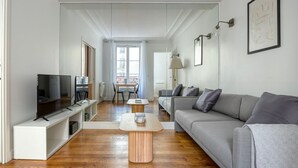 A modern living room with a grey sofa, a wooden coffee table, a TV on a white stand, and a dining area with a table and chairs. - Blueground | Auteuil, concierge, nr Seine & dining (Paris)