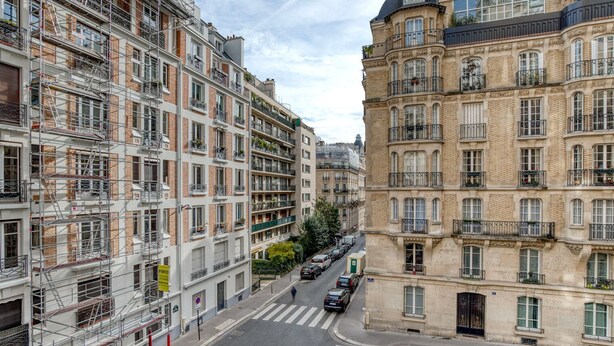 A multi-story urban street with buildings on either side, including a building with scaffolding and another with ornate architecture. - Blueground | Trocadéro, nr shops & Eiffel Tower (Paris)