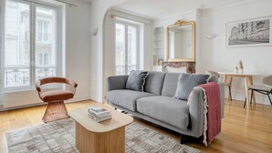 A modern living room with a grey sofa, a wooden coffee table, and a red throw blanket. There is a chair, a small table with two bottles, and a large mirror on the wall. - Blueground | Montmartre, close to shops & cafes (Paris)
