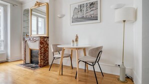 A room with a fireplace, a round table with two chairs, a large mirror, a framed cityscape picture, and a floor lamp. - Blueground | Montmartre, close to shops & cafes (Paris)