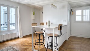 Ein Essbereich mit einem weißen Tisch, vier Hocker, einer Pendelleuchte und Blick auf den Außenbalkon durch ein Fenster.