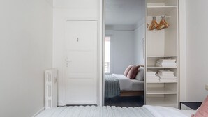 Une chambre avec une porte blanche, un lit recouvert d’une couverture grise, un oreiller rose et une armoire blanche avec des cintres.