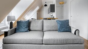 Een moderne woonkamer met een grijze bank, twee blauwe kussens, een lamp en een eethoek op de achtergrond.