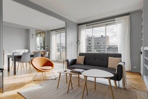 A modern living room with a sofa, a dining table, and chairs. There is a large window with curtains, and the room has a view of buildings outside. - Blueground | Auteuil, balcony & Eiffel Tower view (Paris)