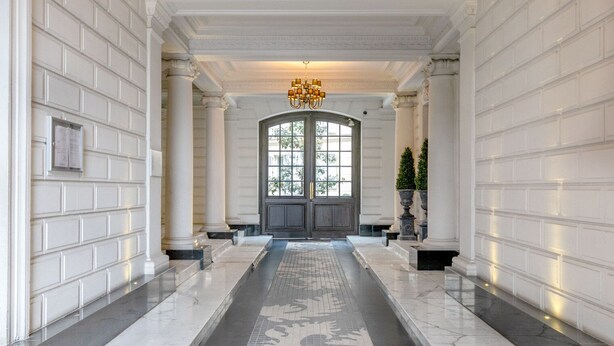Un vaste hall d’entrée aux murs recouverts de panneaux blancs, doté d’un sol en marbre et d’une grande porte à double battant avec un panneau de verre.