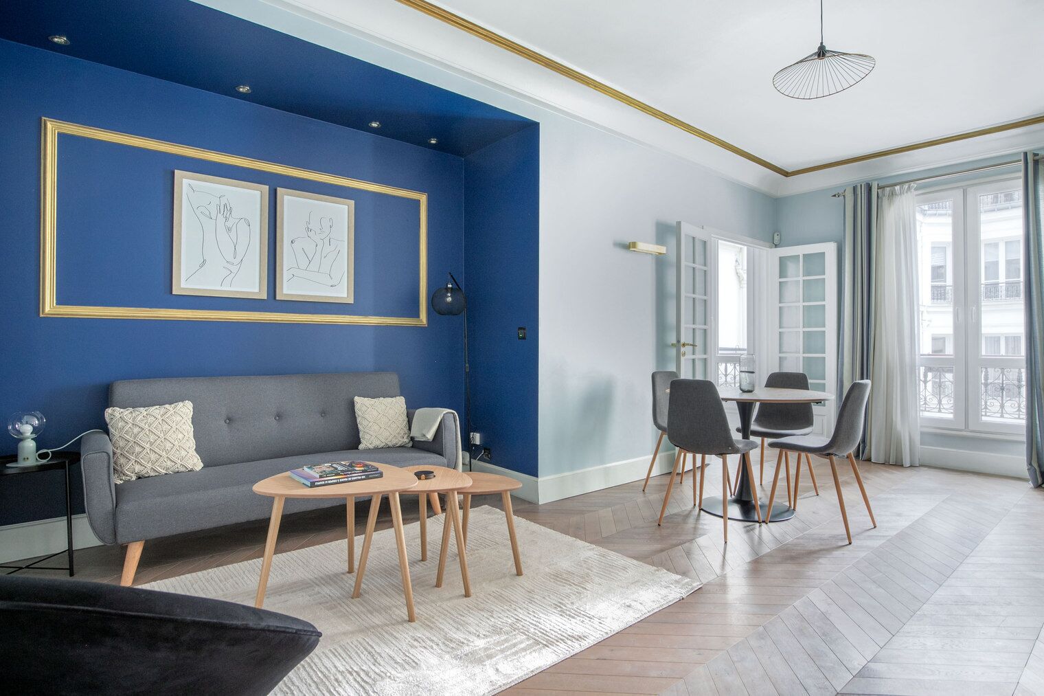 En moderne stue med en blå accentvæg, en grå sofa, et træbord og et spiseområde med et hvidt bord og grå stole.