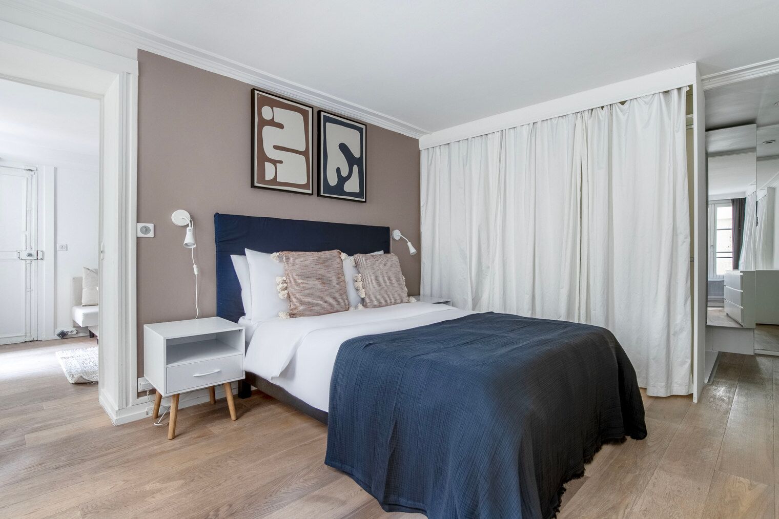 Une chambre à coucher moderne avec un grand lit, une table de chevet et une commode. On y trouve deux lampes fixées au mur et un miroir au-dessus du lit.