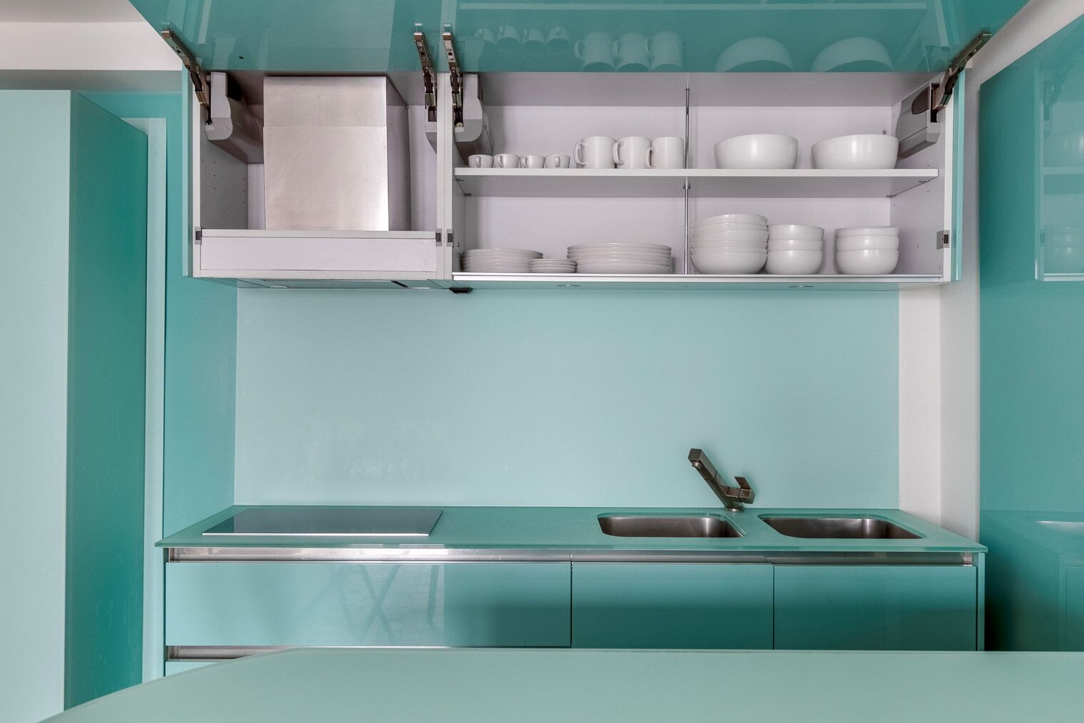 Une cuisine avec des meubles de couleur turquoise, deux éviers en acier inoxydable et de la vaisselle blanche rangée sur des étagères.