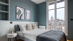 A modern bedroom with a large bed, a small side table, and a view of a cityscape through a large window. - Blueground | Sorbonne, balcony, a/c & great view (Paris)