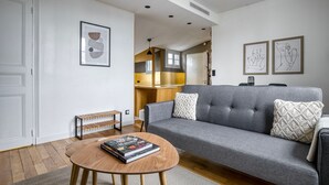 A modern living room with a grey sofa, wooden coffee table, and a bench. There are framed artworks on the wall and a kitchen area in the background. - Blueground | Sorbonne, balcony, a/c & great view (Paris)