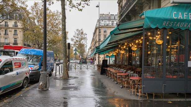 A rainy street with parked cars, a cafe with outdoor seating, and a building with a green awning. - Blueground | Sorbonne, balcony, a/c & great view (Paris)
