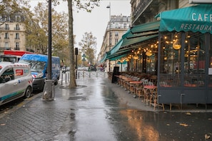 A rainy street with parked cars, a cafe with outdoor seating, and a building with a green awning. - Blueground | Sorbonne, balcony, a/c & great view (Paris)