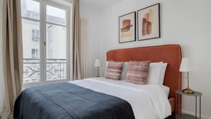 A hotel room with a bed, a nightstand, a lamp, and two framed artworks on the wall. - Blueground | Le Marais, elevator, close to market (Paris)