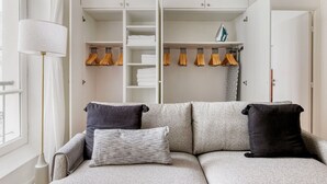 A modern living room with a grey sofa, two black cushions, a white lamp, and a white wardrobe with hanging clothes. - Blueground | Le Marais, elevator, close to market (Paris)