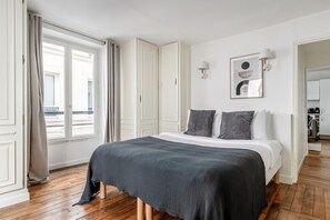 A bedroom with a bed, a window with curtains, a door, and a wall-mounted light fixture. - Blueground | Trocadéro, near Champs Elysées (Paris)
