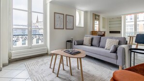 Un salon avec un canapé gris, une table basse ronde et une vue sur le paysage urbain, visible à travers de grandes fenêtres.