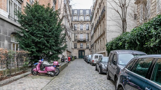 Une rue pavée bordée de voitures et de scooters garés, qui mène à un bâtiment à l’architecture classique.