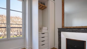 A room with a large window, a wardrobe with multiple drawers, a hanging rack, and a fireplace. - Blueground | Invalides, furnished, nr eiffel tower (Paris)