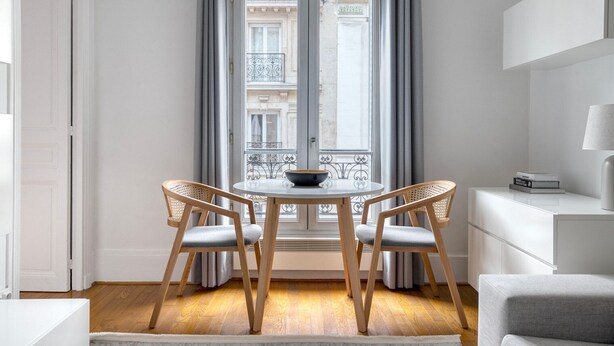 A modern room with a round table, two chairs, a white sofa, a lamp, and a view of buildings through a window. - Blueground | Bastille, nr shopping & dining (Paris)