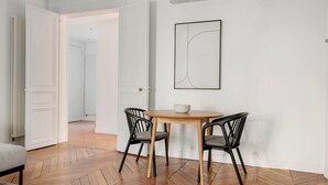 Una habitación con suelo de madera tipo espiga, una mesa redonda de madera con dos sillas negras, una puerta blanca y una obra de arte abstracta enmarcada en la pared.