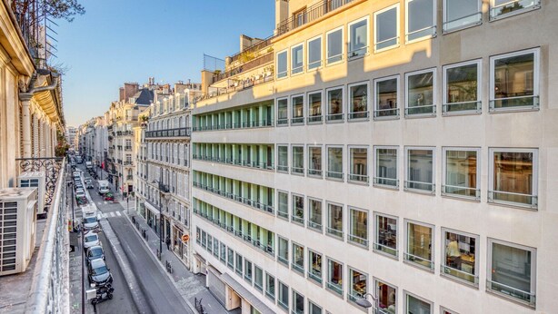 Un edificio de varios pisos con fachada moderna, amplios ventanales y vista a la calle con coches estacionados y una acera.