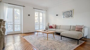 Amplia sala de estar con piso de madera, un sofá color beige, una mesa de centro y ventanas grandes con cortinas.