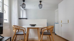 A modern kitchen with two wooden chairs, a white table, and a black bowl. There are two pendant lights, a white cabinet, and a window with curtains. - Blueground | Passy, walk to park & foodie area (Paris)
