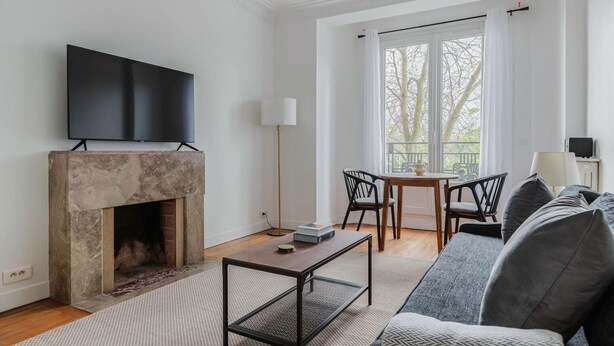 A modern living room with a fireplace, a flat-screen TV mounted above it, a sofa, a coffee table, and a balcony with a view of trees.