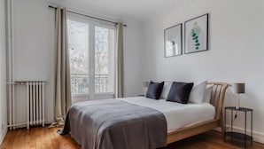 A bedroom with a bed, a radiator, a window with curtains, a bedside table with a lamp, and two framed artworks on the wall.