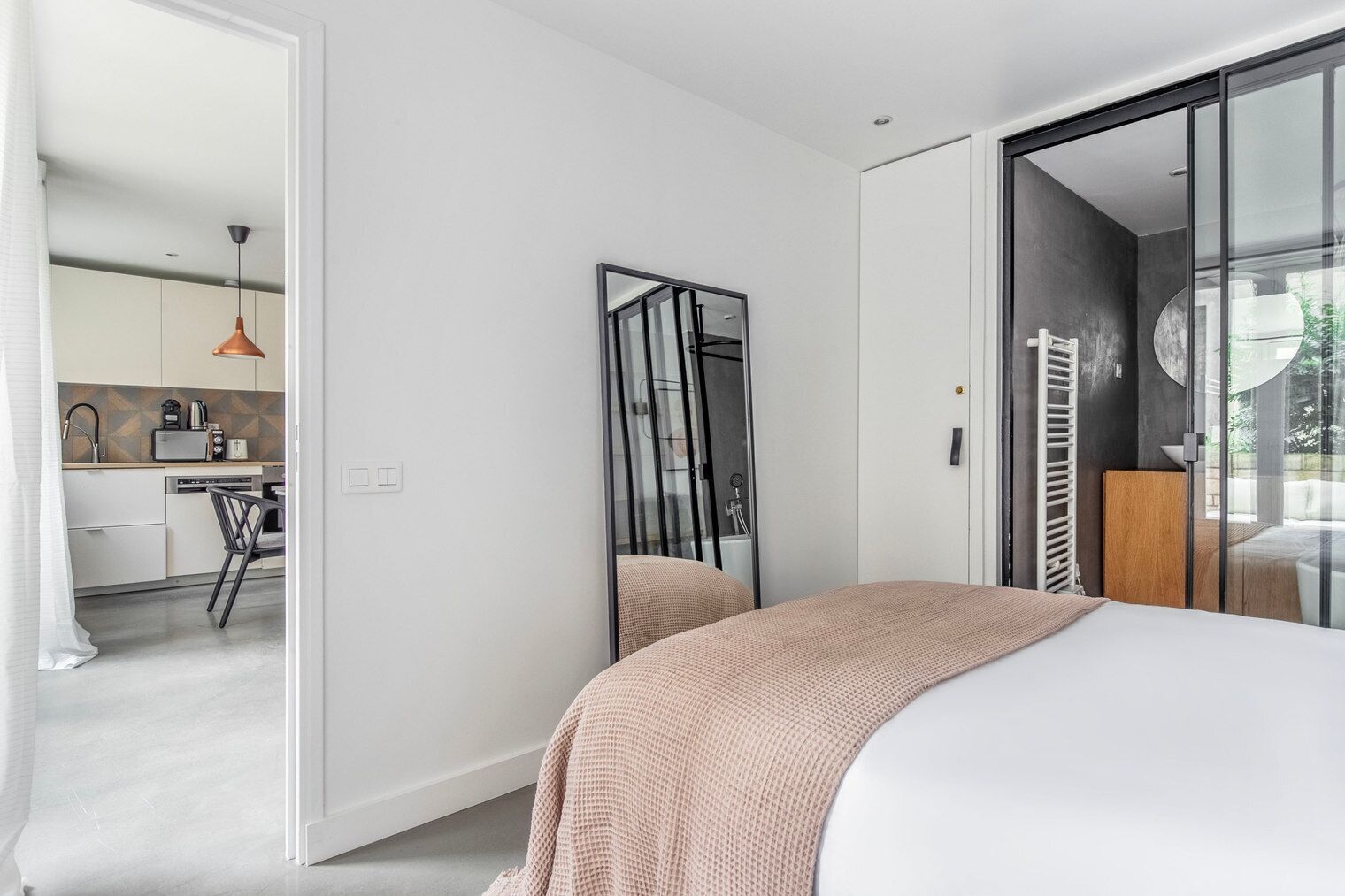 Une chambre à coucher moderne avec un grand lit, une porte en verre donnant sur une salle de bain et une cuisine visible à travers une porte ouverte.