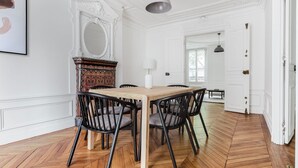 A dining area with a wooden table and chairs, white walls, and a herringbone floor pattern. - Blueground | Jardin du Luxembourg, walk to gardens (Paris)