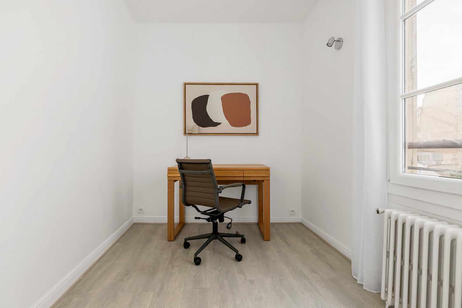 Ett litet, rent rum med ett skrivbord och en kontorsstol i trä, en inramad abstrakt målning och ett fönster med utsikt över byggnader.