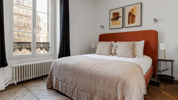 Ein Schlafzimmer mit einem großen Bett, einem Fenster mit Vorhängen, einem Heizkörper und zwei gerahmten Kunstwerken an der Wand.
