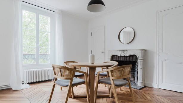 A room with a wooden herringbone floor, a round table with four chairs, a white radiator, a fireplace, and a large window with white curtains. - Blueground | Jardin du Luxembourg, walk to gardens (Paris)