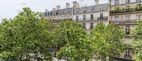 A row of multi-story buildings with balconies, trees in the foreground, and a clear sky.