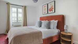 Une chambre à coucher bien rangée, avec un grand lit, une table de chevet dotée d’une lampe et une fenêtre avec des rideaux.