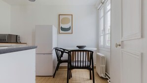 A compact kitchen with a white refrigerator, a microwave, and a small dining table with two chairs. A framed artwork hangs on the wall above the table. - Blueground | Jardin du Luxembourg, near gardens (Paris)