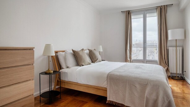 Un dormitorio ordenado con una cama de madera, una mesita de noche con una lámpara y vistas a la ciudad a través de un ventanal grande.