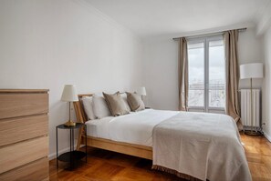 Ein ordentlich eingerichtetes Schlafzimmer mit einem Holzbett, einem Nachttisch mit Lampe und Blick auf die Stadt durch ein großes Fenster.