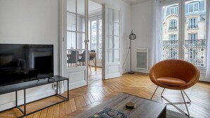 A living room with a TV, a brown chair, a coffee table, and a view of a balcony with buildings. - Blueground | Champ de Mars, walk to Eiffel Tower (Paris)